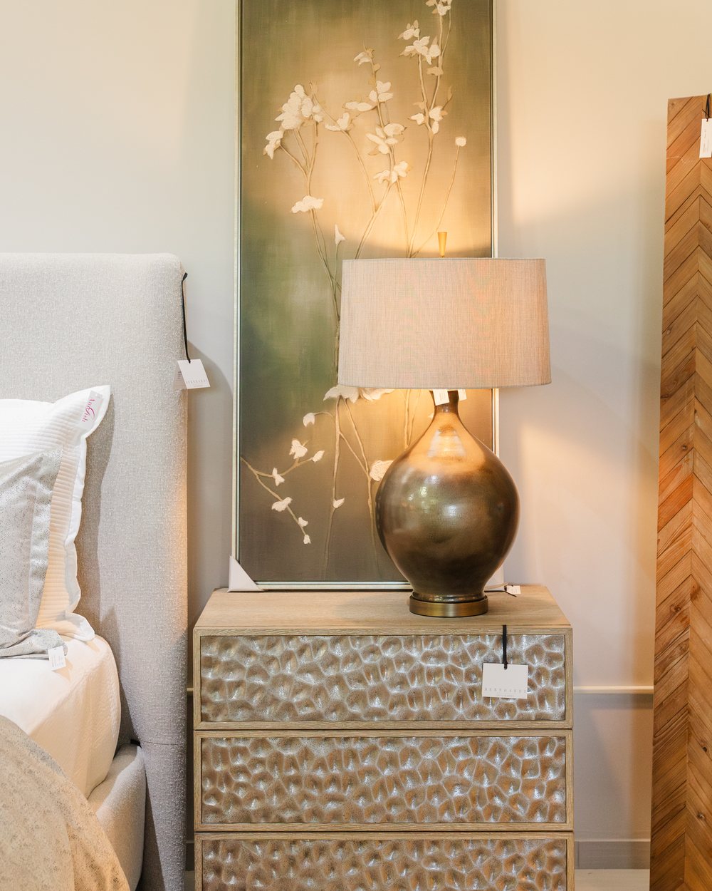A hammered-front nightstand with a bronze lamp beneath a lit botanical canvas
