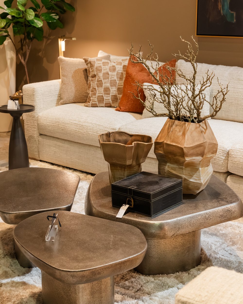 A cream sofa with rust and patterned pillows above gold sculptural cocktail tables