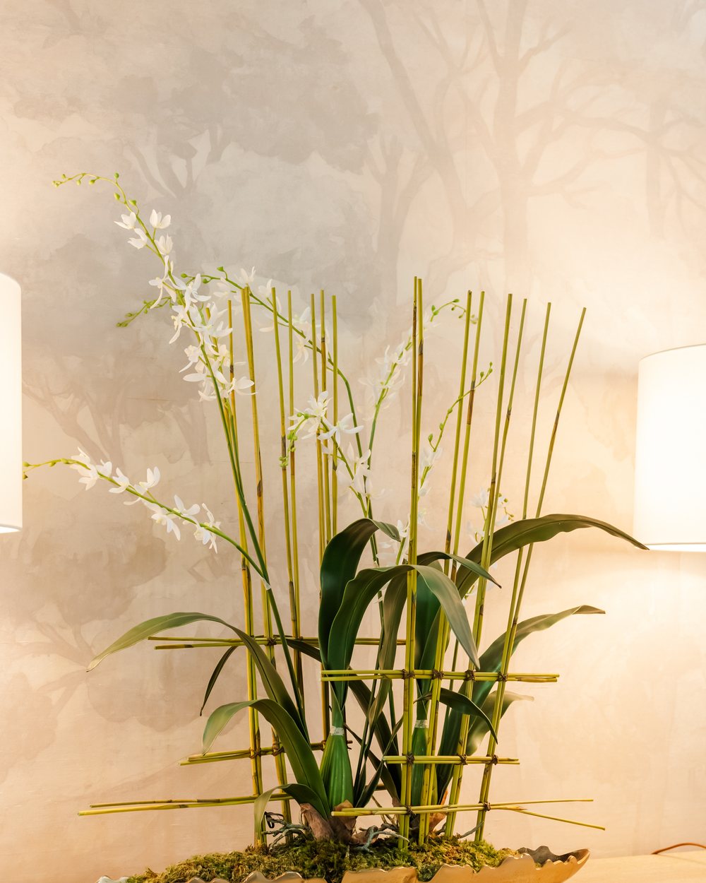 A white orchid and reed arrangement in a gold bowl against a soft landscape mural