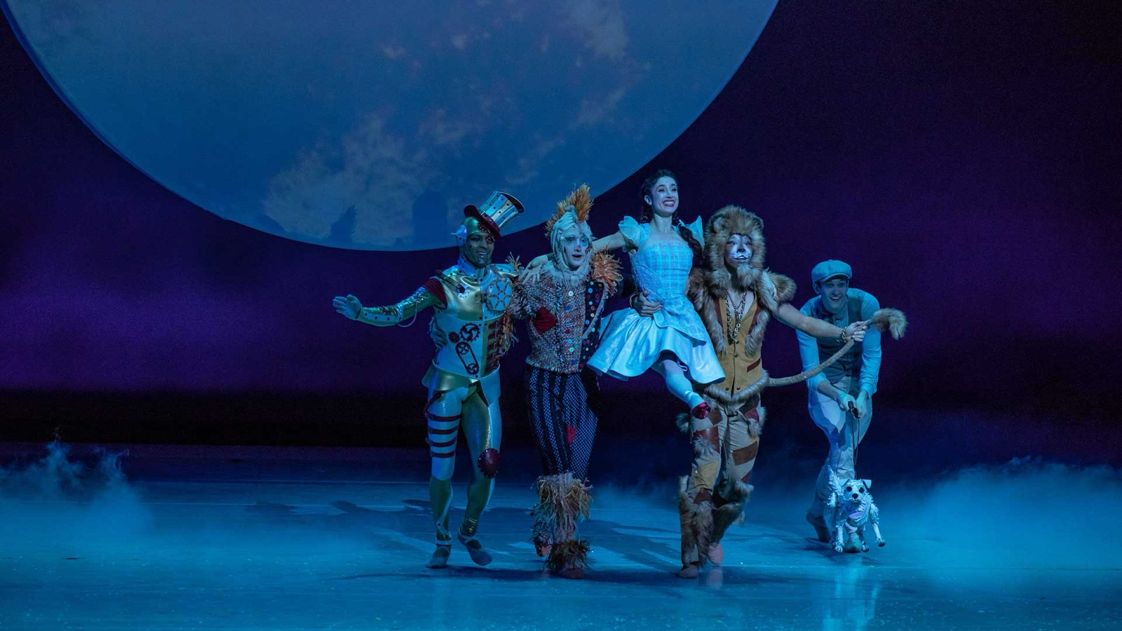 Dorothy, the Tin Man, the Scarecrow, and the Lion walk arm in arm under a full moon with Toto alongside, in Kansas City Ballet's The Wizard of Oz