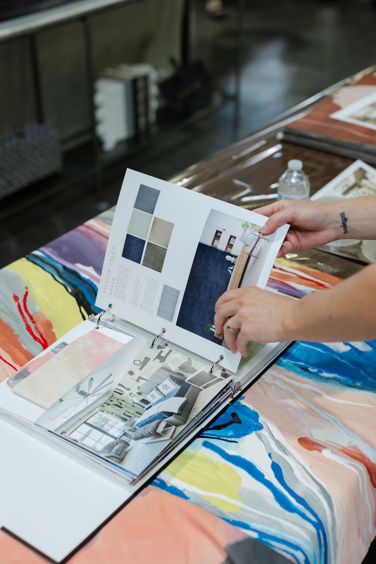 Hands leafing through the open Porter Teleo lookbook, mood boards and material swatches on view, atop a table draped in the studio's hand-painted textile