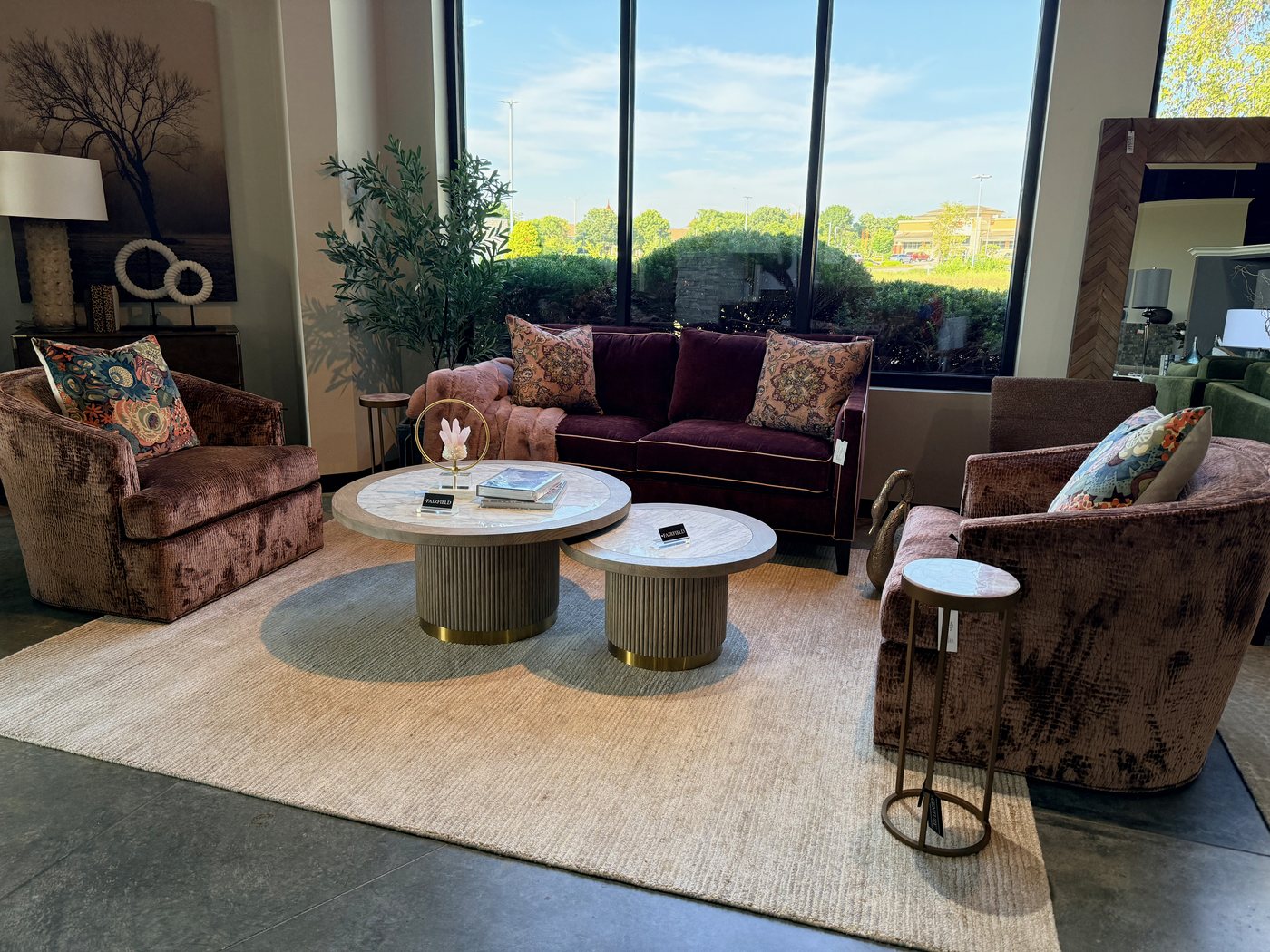 Two rose crushed-velvet swivel chairs angled toward each other across a pair of round fluted cocktail tables, with a small drink table beside one chair and a plum velvet sofa under the windows behind them, in the Seville Home showroom.
