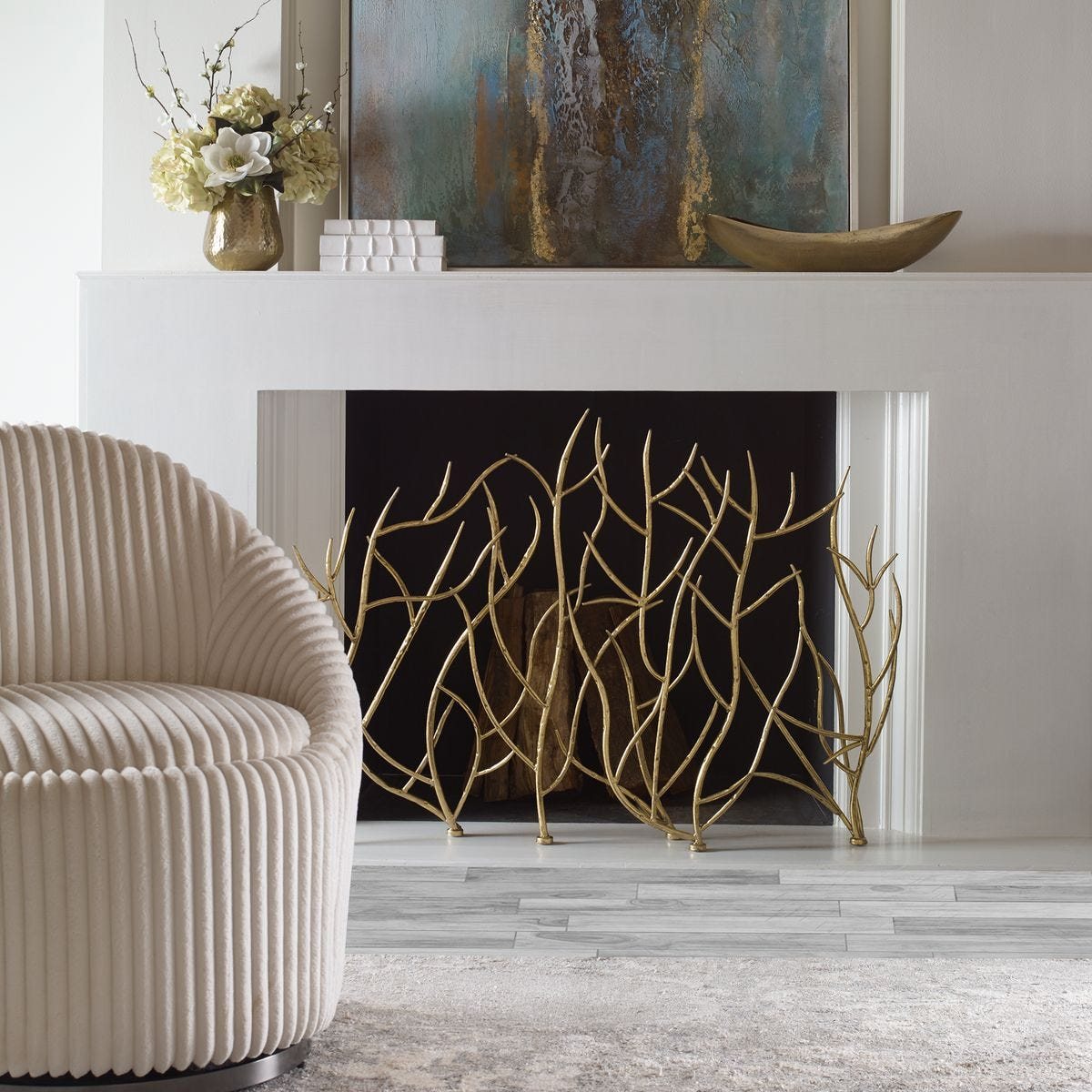 A white mantel with abstract art leaned against the wall, a brass vase of white magnolias and hydrangea at the left end, a short stack of white boxes beside it, and a low brass bowl at the right.