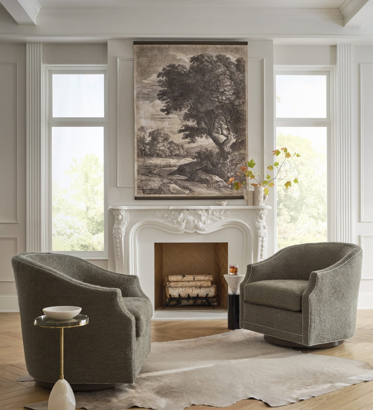 A carved white mantel beneath a large landscape engraving, with a small arrangement of autumn branches at one end of the shelf and a pair of upholstered swivel chairs facing the fireplace.
