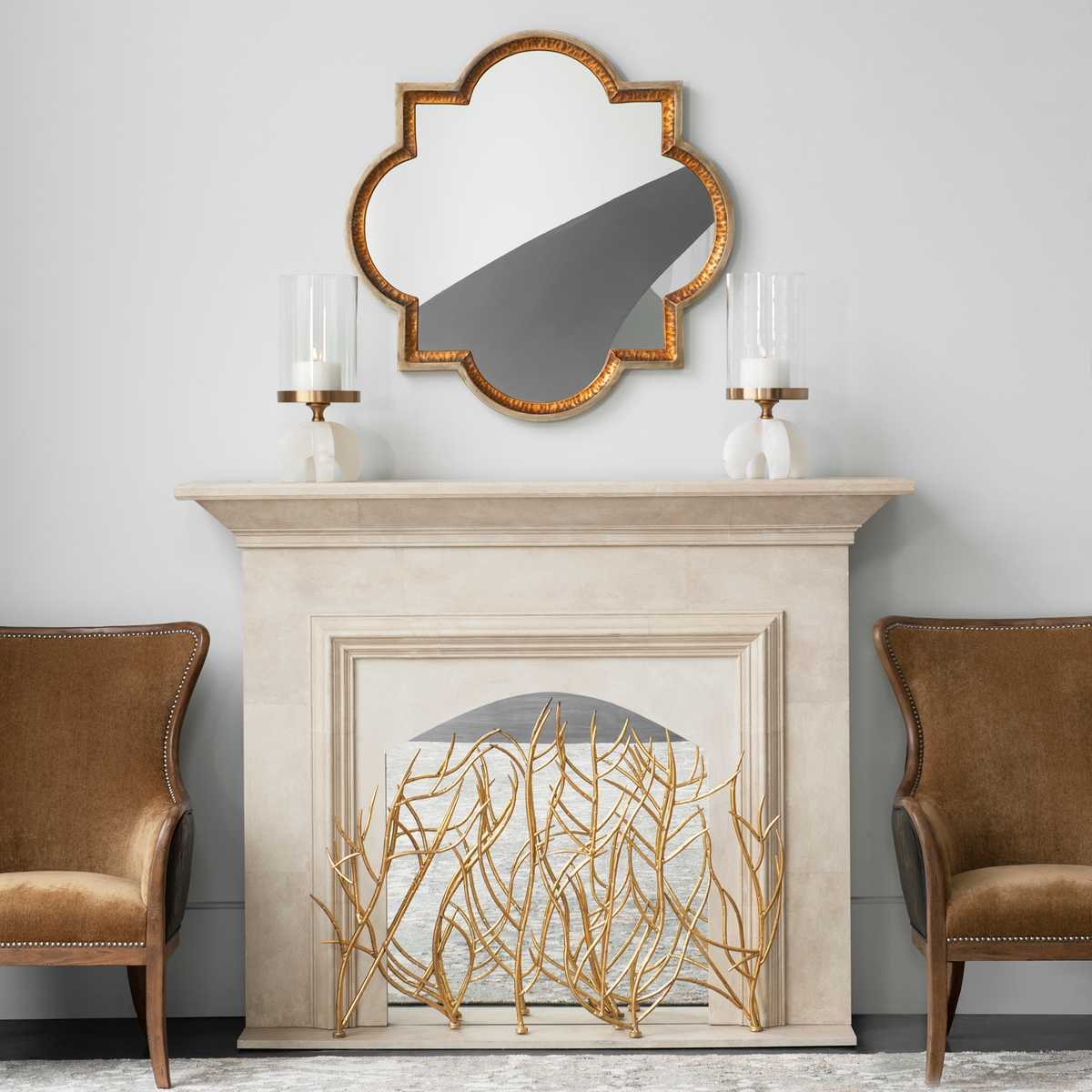 A cream cast-stone mantel with a gold quatrefoil mirror centered above it, a matched glass hurricane on a stand at each end, and a gold branch fireplace screen in the opening.
