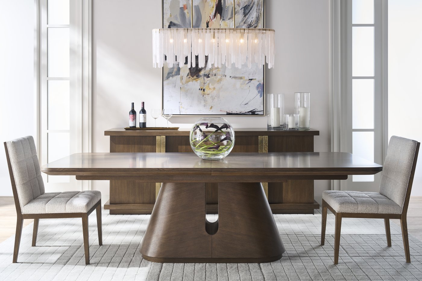The Century Carino dining table in Cocoa on its sculpted pedestal, with a low glass bowl of calla lilies at the center and a sideboard set as a bar behind it.