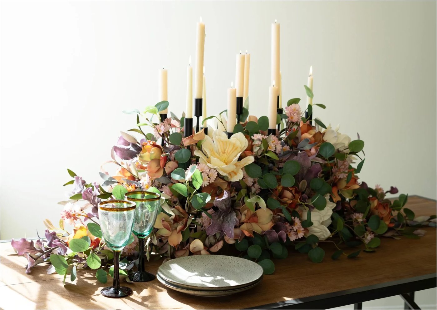A black iron candelabra of cream taper candles nested in a long, low arrangement of faux hydrangea, magnolia, and eucalyptus on a wood dining table, with stacked plates and green-rimmed goblets at one end.