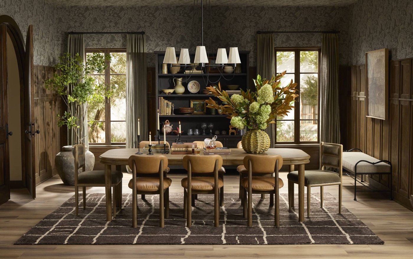 The Four Hands Florent oak dining table in a wood-paneled dining room, styled with taper candles, books, and a tall arrangement of hydrangea and magnolia leaves, with leather and velvet dining chairs.