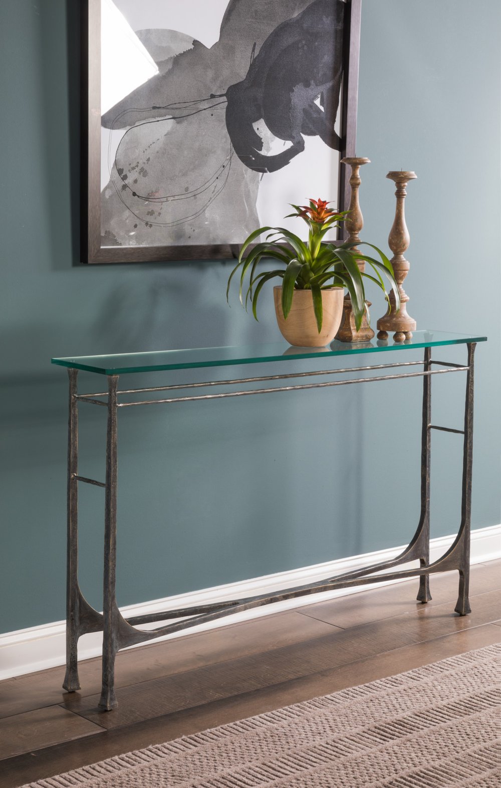 A slim forged-iron console with a glass top against a teal wall, holding a potted bromeliad and two turned wooden candlesticks of different heights beneath a framed abstract print.