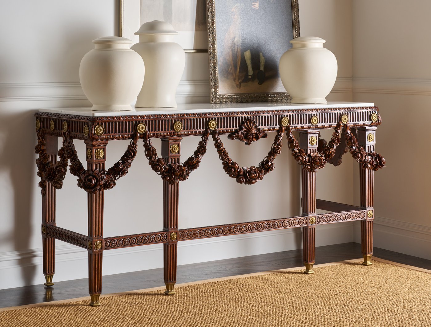 A carved mahogany console with a white marble top and rose garland swags along its frieze, holding three cream ceramic urns, two clustered and one apart, in front of a leaning framed portrait.