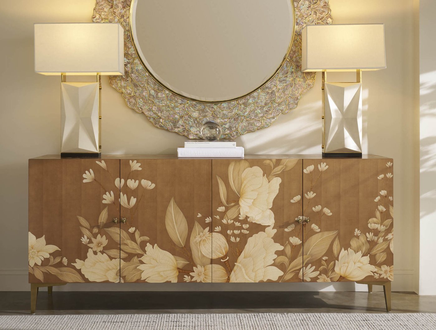 A Theodore Alexander sideboard with floral inlay across four doors, a round shell-framed mirror centered above it, and a matched pair of lamps at its ends.
