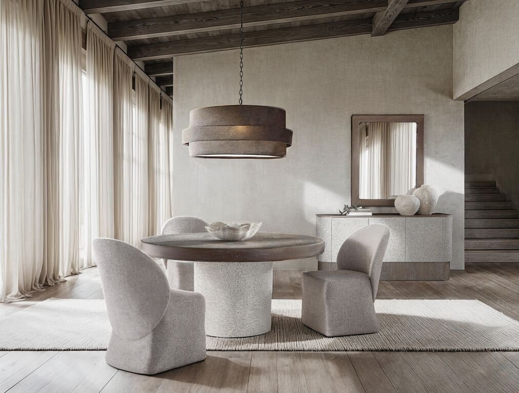 A dining room with a round pedestal table and upholstered chairs; behind them, a textured ivory buffet sits beneath a wood-framed mirror, styled with two pottery vessels and a small object at one end.