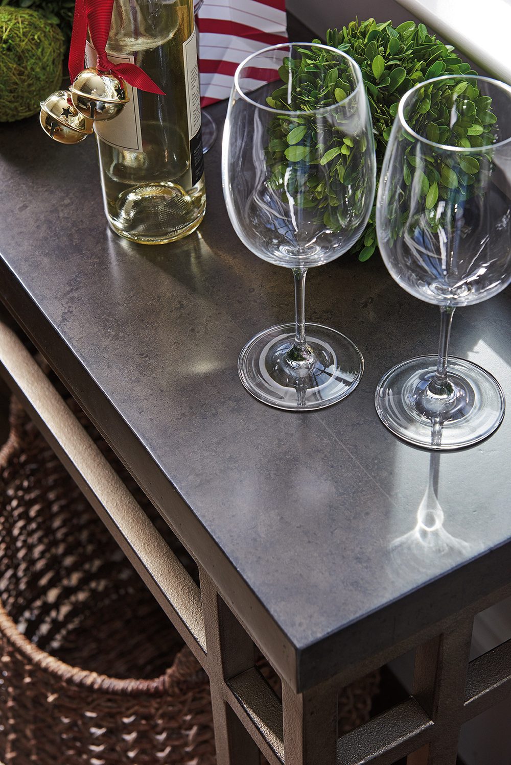 The corner of a dark-topped Lexington Twin Lakes sideboard dressed for the holidays with a boxwood topiary, wine glasses, and a bottle tied with a red ribbon and gold jingle bells.