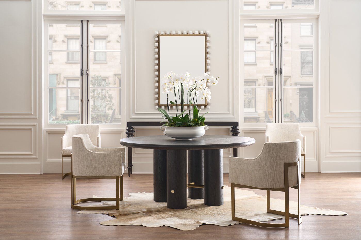 The Century Cadence 64-inch round dining table in Mocha oak on four columns with a brass stretcher, set with four upholstered armchairs beneath a beaded mirror.