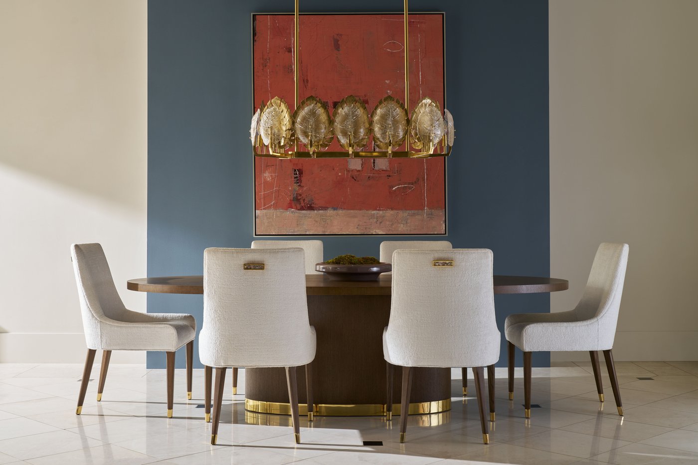 The Theodore Alexander Judith Leiber Couture oval dining table in Mink oak with a brass-banded plinth base, set with six cream upholstered side chairs beneath a brass leaf chandelier.