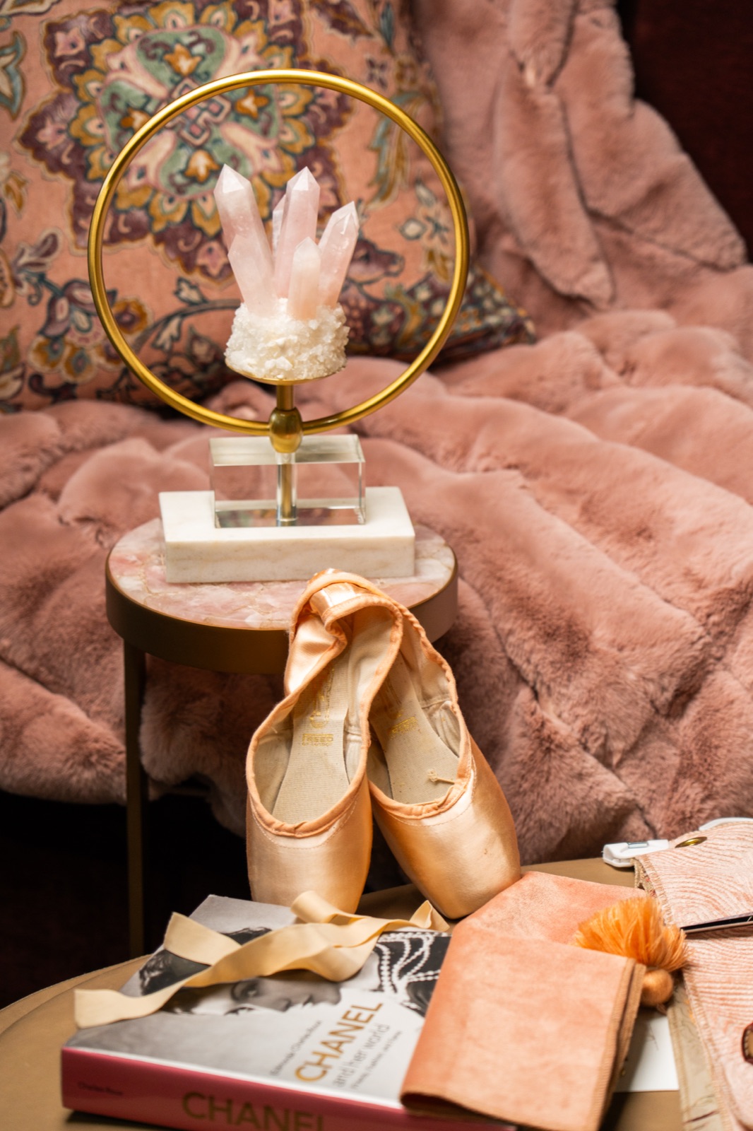 Blush pointe shoes beside a faux-fur throw, a rose quartz accent and a stack of design books