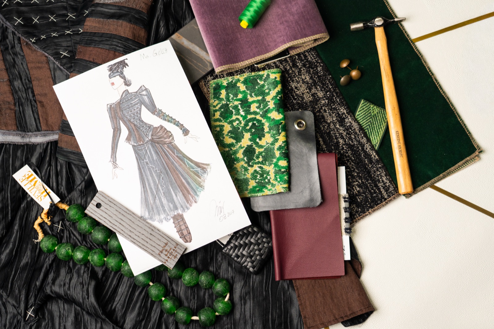 An Emerald City costume sketch laid over green velvet, damask, leather and a strand of green beads