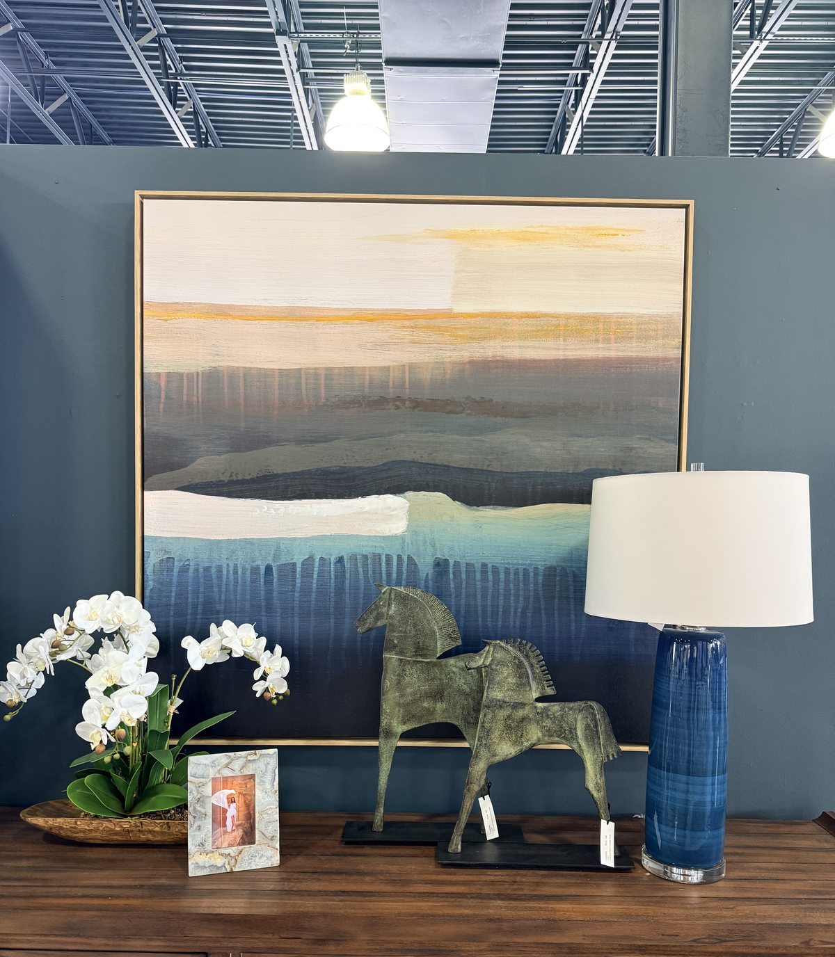A carved antique chest styled with white orchids, a bronze horse, and a blue ombre lamp beneath a sunset landscape painting
