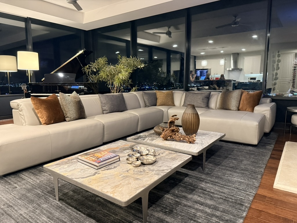 The same great room, styled and at rest: a cream leather sectional layered with cognac and charcoal pillows beside a grand piano and a wall of city lights