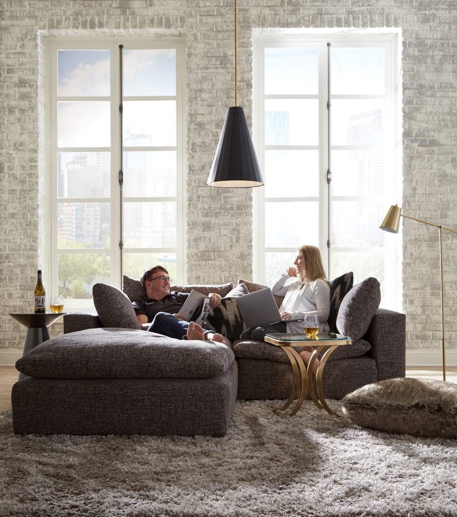 A couple relaxing together on a deep charcoal Huntington House sectional in a sunlit loft, laughing over a laptop and a glass of wine