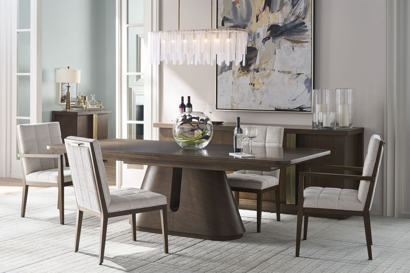 The Century Carino dining table set with upholstered chairs beneath a linear crystal chandelier, a walnut sideboard behind it.