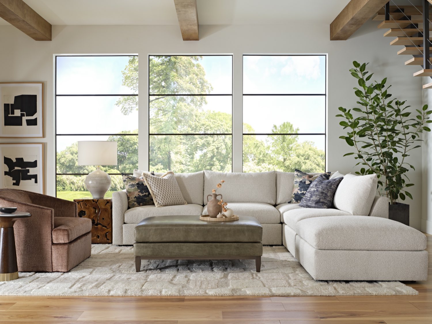 A bright, high-ceilinged living room styled around a cream bouclé Massoud sectional and a leather ottoman, with a driftwood side table, tall windows, and a floating staircase.